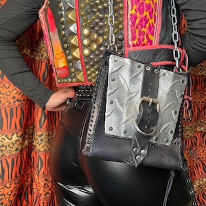 Pu&ograve; includere: Una borsa a tracolla in pelle nera con un pannello in metallo argentato e una fibbia nera. La borsa ha una lunga tracolla a catena e un dettaglio a frangia.
