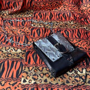 Pu&ograve; includere: Una borsa in pelle nera con un pannello in metallo argentato e una fibbia argentata. La borsa ha una tracolla a catena e si trova su un tessuto a motivi.