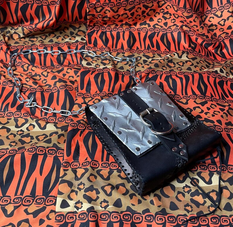 Pu&ograve; includere: Una borsa in pelle nera con un pannello in metallo argentato e una fibbia argentata. La borsa ha una tracolla a catena e si trova su un tessuto a motivi.