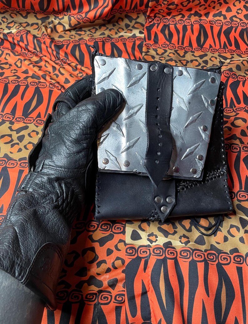 Pu&ograve; includere: Un portafoglio in pelle nera con un design a piastrelle di diamante argentate e un cinturino in pelle nera. Il portafoglio ha rivetti argentati ed &egrave; tenuto in una mano che indossa un guanto in pelle nero.