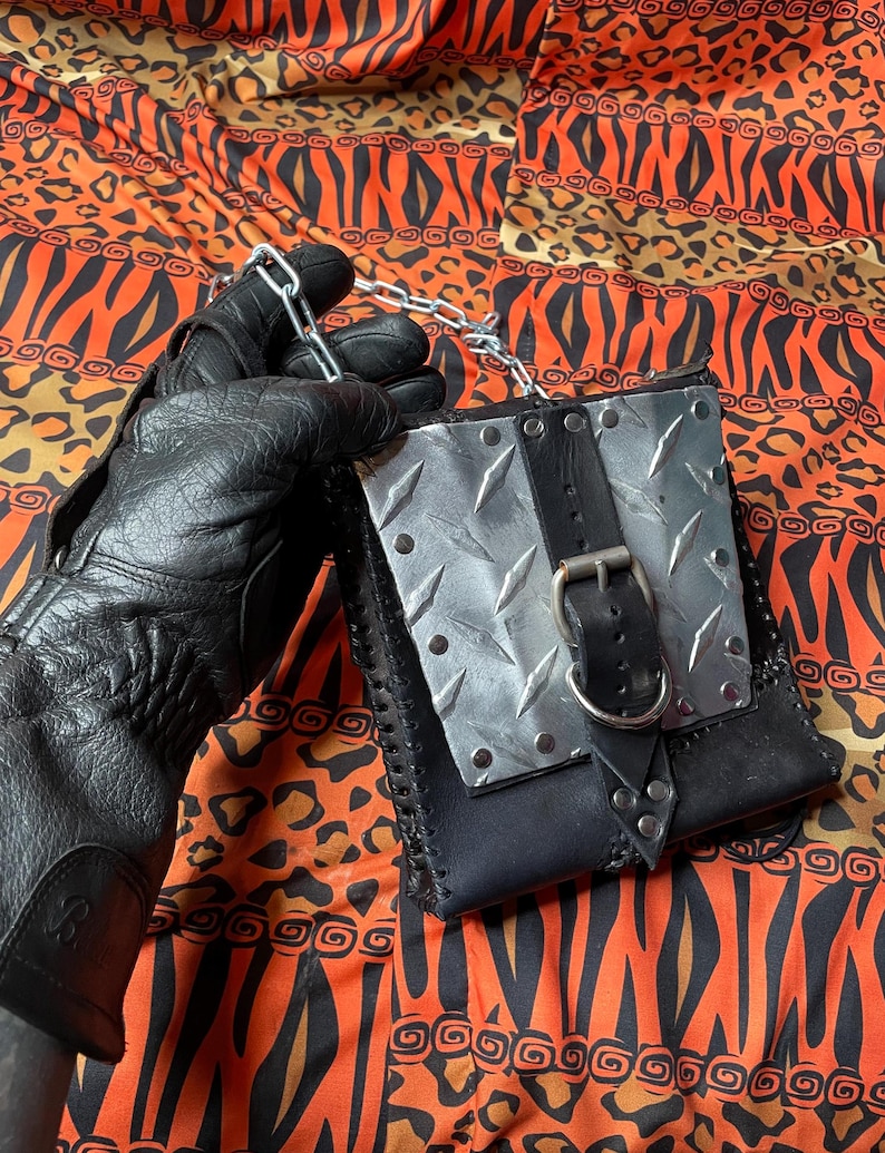 Pu&ograve; includere: Una borsa in pelle nera con un pannello in metallo argentato e una fibbia nera. La borsa ha una tracolla a catena ed &egrave; decorata con borchie argentate.