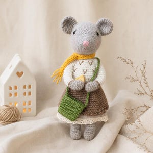 Könnte beinhalten: Eine gehäkelte Mauspuppe mit grauem Fell, einer rosa Nase und schwarzen Augen. Sie trägt einen weißen Pullover mit braunen V-förmigen Stickereien, einen braunen Rock mit weißer Spitzenborte, einen gelben Schal und eine kleine grüne Umhängetasche.