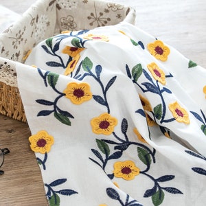 May include: White fabric with embroidered yellow flowers and green leaves on a vine. The fabric is folded and displayed in a wicker basket.