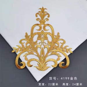 May include: A gold-colored embroidered applique with an ornate floral design. The applique is approximately 8.66 inches wide and 9.45 inches high. It is displayed on a white fabric background.