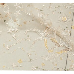 May include: Ivory-colored fabric with embroidered floral and star patterns. The fabric is adorned with sequins and delicate, flowing designs. The embroidery features a mix of cream and gold threads, creating a subtle, elegant texture. Ideal for bridal or formal wear.