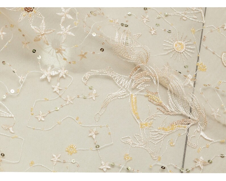 May include: Ivory-colored fabric with embroidered floral and star patterns. The fabric is adorned with sequins and delicate, flowing designs. The embroidery features a mix of cream and gold threads, creating a subtle, elegant texture. Ideal for bridal or formal wear.
