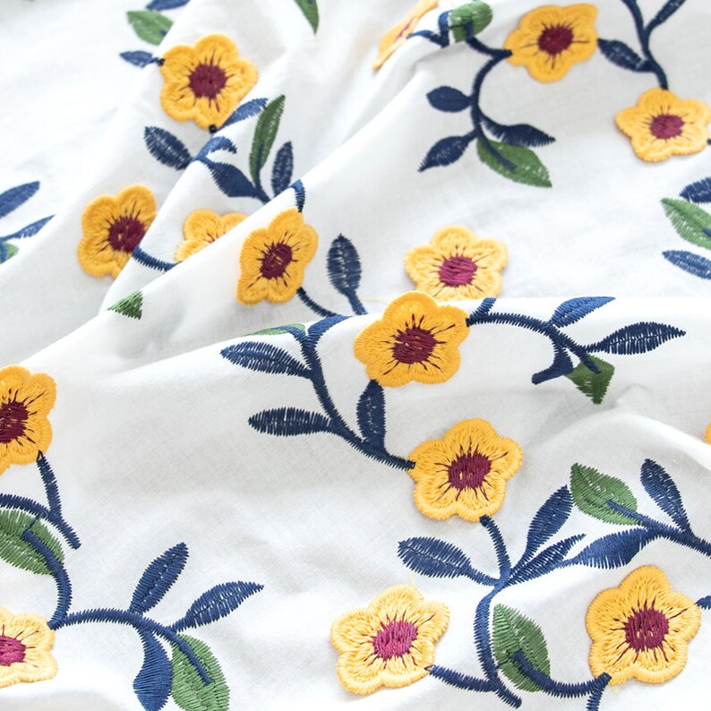May include: White fabric with yellow embroidered flowers and blue and green leaves. The flowers have a burgundy center.