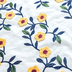 May include: White fabric with embroidered yellow flowers with maroon centers and dark blue and green stems.