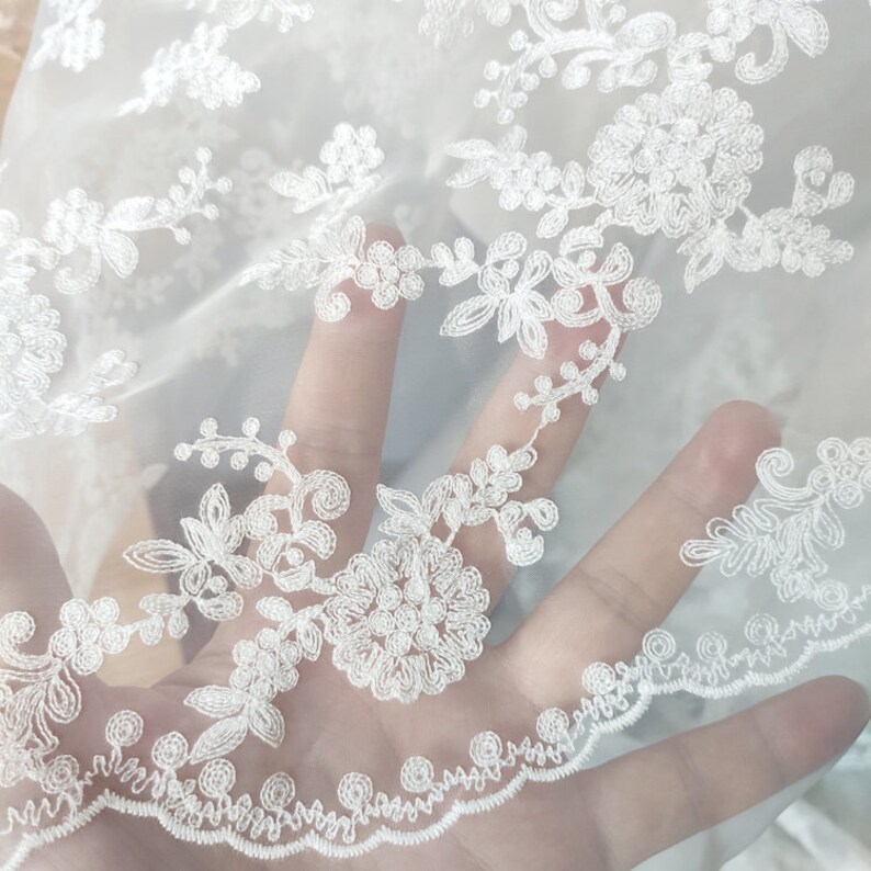May include: White sheer fabric with delicate floral embroidery. The fabric has a soft, flowing texture and is perfect for creating elegant garments or accessories.