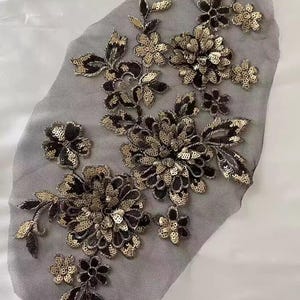 May include: A black and gold sequined floral applique on grey mesh. The design features multiple flower clusters and leaf accents. The sequins provide a shimmering effect, ideal for embellishing garments or crafts.