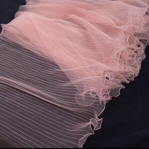 Peut inclure: Gros plan sur un tissu rose p&acirc;le d&eacute;licat, &agrave; texture pliss&eacute;e. Le tissu est transparent et pr&eacute;sente un bord festonn&eacute;. Le mat&eacute;riau semble l&eacute;ger et fluide, adapt&eacute; &agrave; l'habillement ou &agrave; des fins d&eacute;coratives.