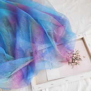 Könnte beinhalten: Ein Stück schimmernder blauer, lila und rosa Tüllstoff mit glitzernder Oberfläche. Der Stoff ist durchsichtig und hat eine weiche, fließende Textur.