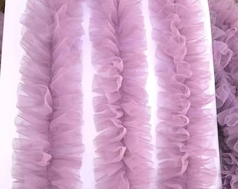 Può includere: Primo piano di tessuto di tulle lavanda disposto a strisce verticali. Il tessuto è arricciato, creando un effetto strutturato e stratificato. Il tulle è un materiale morbido e trasparente, ideale per l'artigianato o la decorazione. Lo sfondo è bianco.