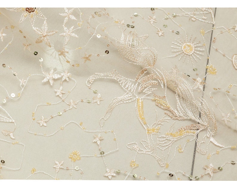 May include: Ivory-colored fabric with embroidered floral and star patterns, accented with sequins. The delicate design features various star and flower shapes, creating a textured, elegant look. The fabric is suitable for apparel and decorative projects.
