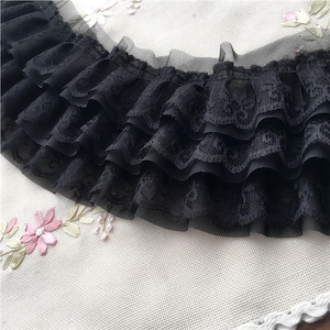 May include: Black lace trim with multiple ruffled layers. The trim has a delicate, floral lace pattern and is layered with a sheer, ruffled fabric. The trim is on a white background with embroidered flowers.