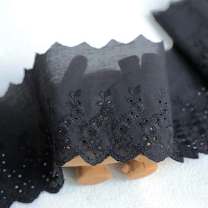 Pode incluir: Acabamento em renda de olhal preta com borda ondulada. A guarnição apresenta bordados florais intrincados e pequenos recortes circulares. O tecido é semi-transparente, ideal para costura e projetos de artesanato.