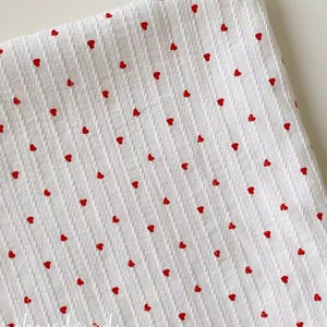 May include: White fabric featuring a textured, vertically striped design. Small, evenly spaced red hearts are scattered across the surface. The fabric appears to be a soft, woven material, suitable for various craft projects.