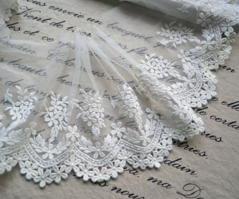 May include: White lace trim featuring a delicate floral design. The lace is made from a soft, sheer fabric and has a scalloped edge.