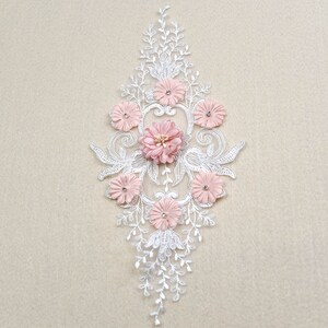 Könnte beinhalten: Eine zarte weiße Spitzenapplikation mit rosa Stoffblumen. Die Applikation hat ein florales Design mit filigranen Details und einer zentralen Blumenansammlung.