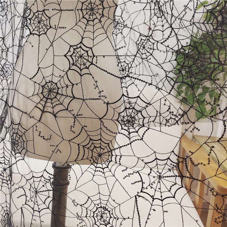 May include: A sheer black fabric featuring an intricate embroidered spider web pattern. The design consists of multiple interconnected webs with delicate black thread detailing and small, reflective black sequins stitched along the web lines.