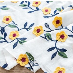May include: White fabric with yellow embroidered flowers and blue leaves. The fabric is folded in half, showing the pattern on both sides.