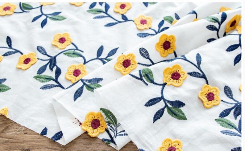 May include: White fabric with yellow embroidered flowers and blue leaves. The fabric is folded in half, showing the pattern on both sides.