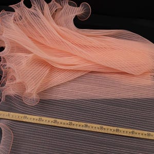 Peut inclure: Un tissu p&ecirc;che pli&eacute; avec un design pliss&eacute; et un bord festonn&eacute;. Une r&egrave;gle en bois avec des mesures en centim&egrave;tres est plac&eacute;e sur le tissu, indiquant des mesures allant jusqu'&agrave; 55 pouces (environ 140 cm).