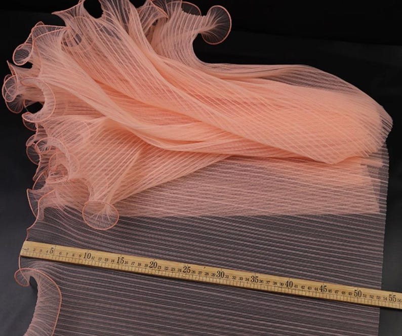 Peut inclure: Un tissu p&ecirc;che pli&eacute; avec un design pliss&eacute; et un bord festonn&eacute;. Une r&egrave;gle en bois avec des mesures en centim&egrave;tres est plac&eacute;e sur le tissu, indiquant des mesures allant jusqu'&agrave; 55 pouces (environ 140 cm).