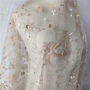 May include: Beige tulle fabric with gold embroidered stars, a sun, and a unicorn. The fabric is embellished with sequins.