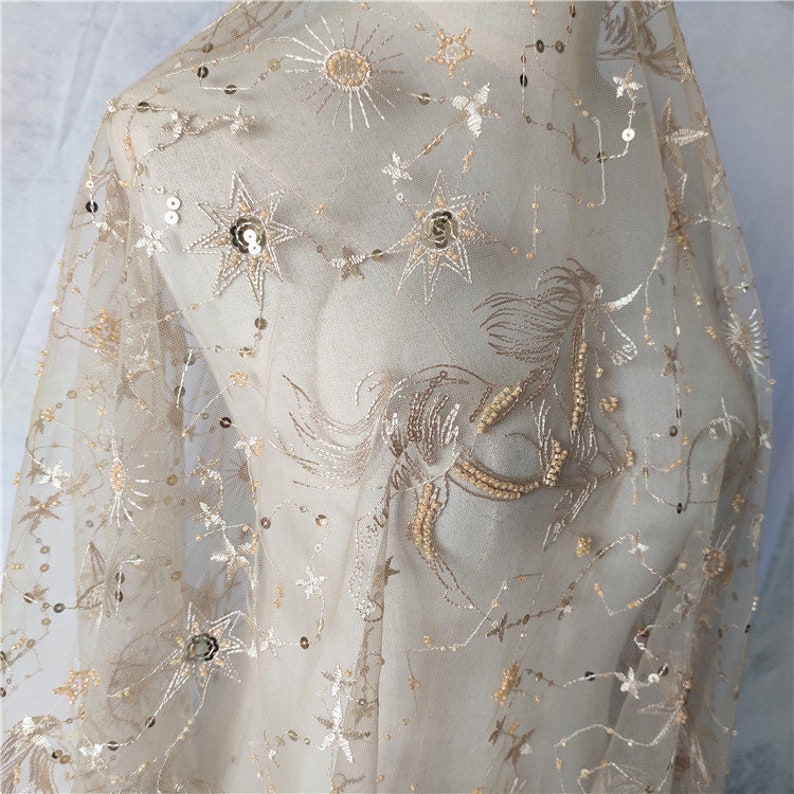 May include: Beige tulle fabric with gold embroidered stars, a sun, and a unicorn. The fabric is embellished with sequins.