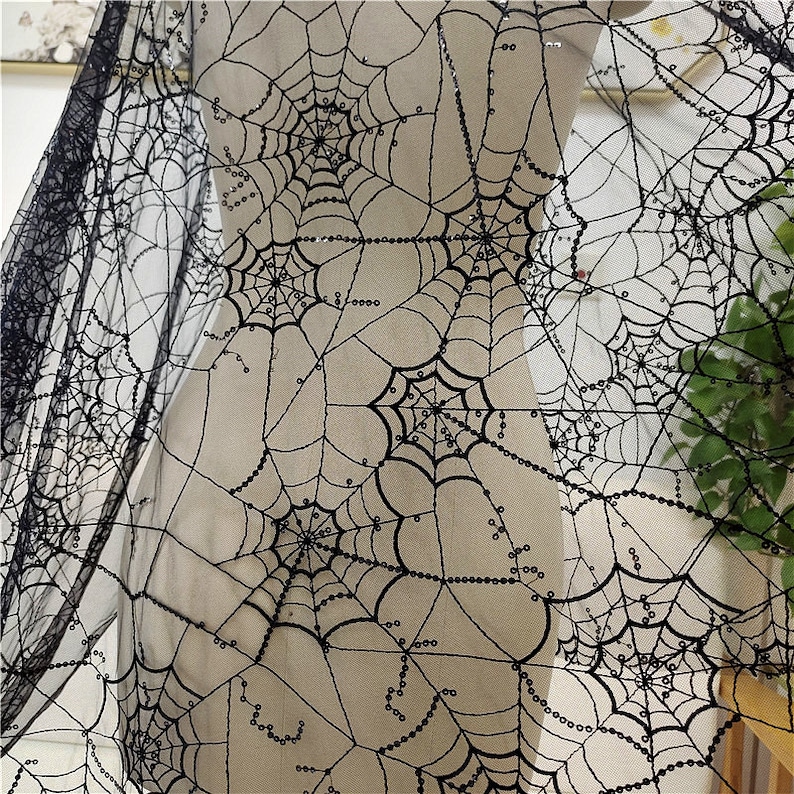 May include: A sheer black fabric featuring an intricate embroidered spider web pattern. The design consists of multiple interconnected webs with black thread and small, reflective black sequins stitched along the lines of the web structure.