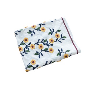 May include: White fabric with embroidered yellow flowers and dark blue stems. The fabric is folded and has a burgundy border.
