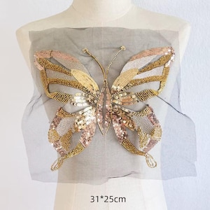 Könnte beinhalten: Ein schmetterlingsförmiger Aufnäher aus goldenen, bronzefarbenen und silbernen Pailletten auf einem transparenten grauen Netzstoff. Das Design zeigt detaillierte Perlenarbeiten an den Flügeln und Fühlern, mit den Maßen 31 mal 25 cm darunter.