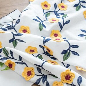 May include: White fabric with yellow embroidered flowers and green and blue leaves. The fabric is folded and has a textured surface.