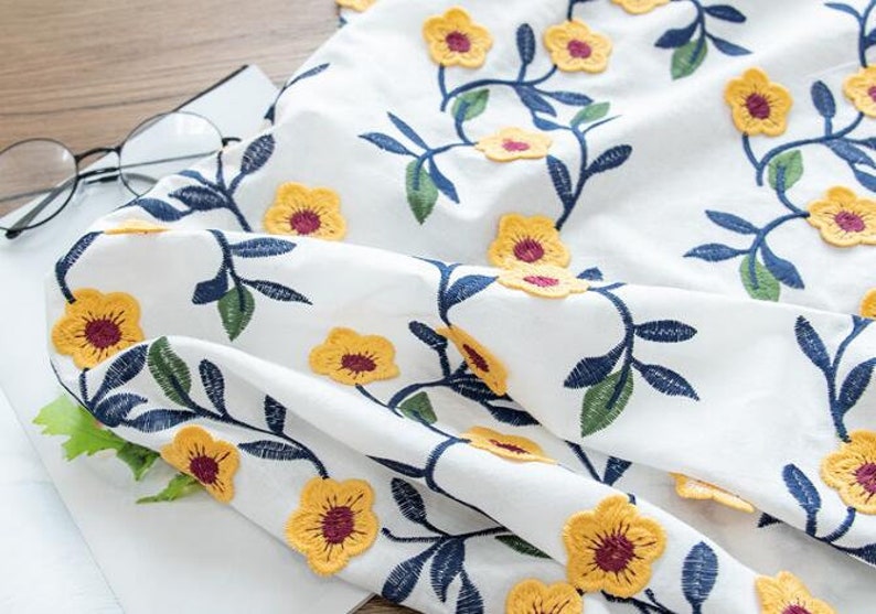 May include: White fabric with yellow embroidered flowers and green and blue leaves. The fabric is folded and has a textured surface.