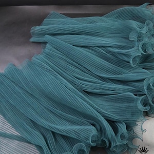 May include: Close-up of teal pleated fabric. The fabric is arranged in flowing, layered folds, creating a textured and dynamic visual effect. The edges of the fabric have a ruffled trim. The fabric is a teal colour.