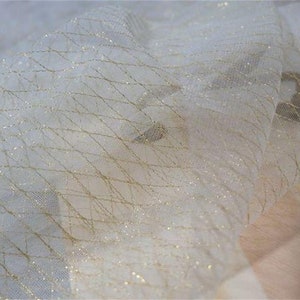 Op de afbeelding: Close-up van een crèmekleurige stof met een ruitpatroon gemaakt door gouden draden. De stof heeft een delicate, transparante uitstraling, met een subtiele glans. Een schaar is zichtbaar in de hoek.