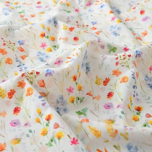 May include: White fabric with a watercolour floral design. The pattern showcases a variety of colourful wildflowers in shades of orange, yellow, pink, blue, and purple. The fabric is gently folded, showing the texture and detail of the print.