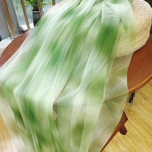 May include: Sheer green and white fabric draped over a woven basket. The fabric has a gradient effect, transitioning from a deep green to a soft white. The fabric is lightweight and appears to be tulle or a similar material, suitable for decorative purposes.