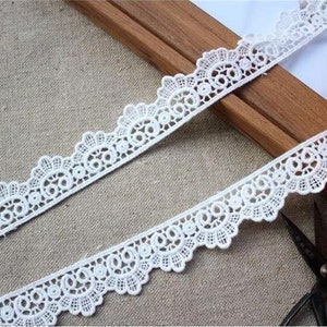 May include: White lace trim with a scalloped edge and delicate floral pattern. The lace is shown on a wooden surface with a pair of scissors.