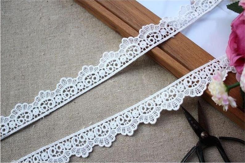 May include: White lace trim with a scalloped edge and delicate floral pattern. The lace is shown on a wooden surface with a pair of scissors.