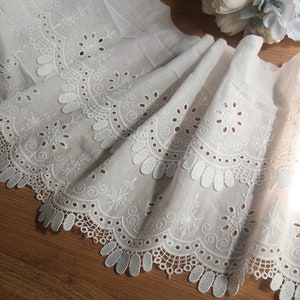 May include: White cotton lace trim with a scalloped edge and delicate eyelet details. The lace has a repeating pattern of circles and swirls.