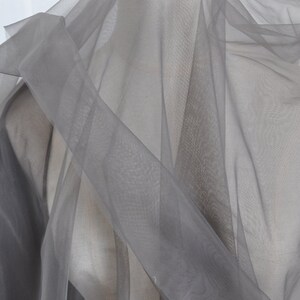 Pu&ograve; includere: Tessuto trasparente grigio con una texture morbida e delicata. Il tessuto &egrave; drappeggiato e piegato, creando un aspetto fluido ed etereo.