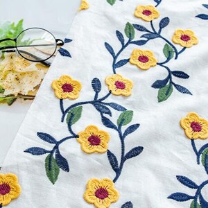 May include: White fabric with embroidered yellow flowers and blue stems. The flowers have a burgundy center.