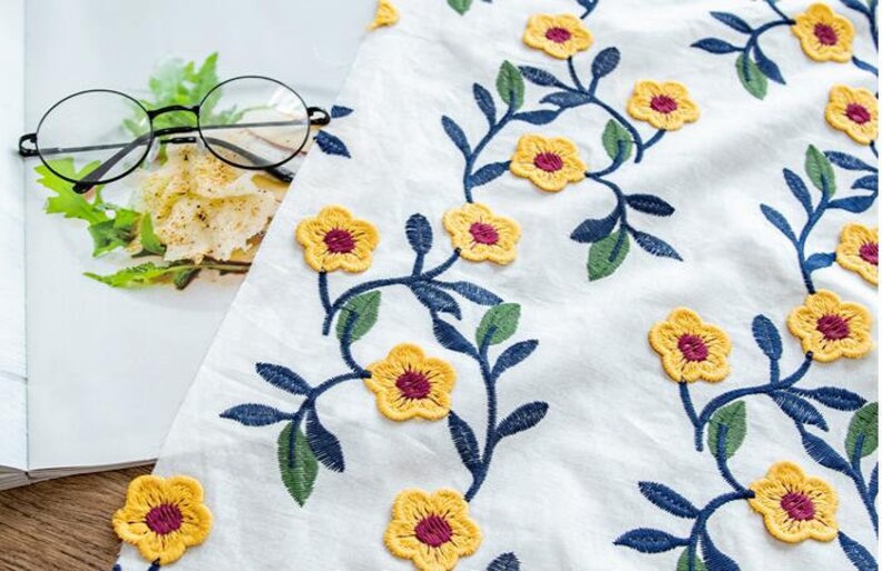 May include: White fabric with embroidered yellow flowers and blue stems. The flowers have a burgundy center.