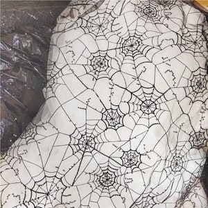 May include: A sheer white fabric featuring an embroidered black spiderweb pattern. The web design is accented with small, reflective black sequins that follow the lines of the webbing, creating a textured and shimmering appearance across the material.