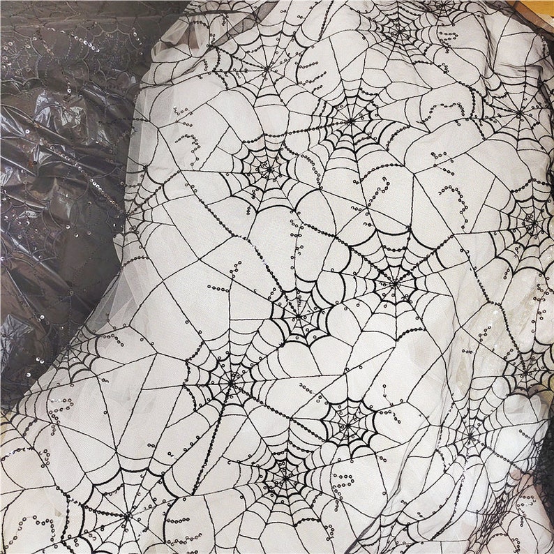 May include: A sheer white fabric featuring an embroidered black spiderweb pattern. The web design is accented with small, reflective black sequins that follow the lines of the webbing, creating a textured and shimmering appearance across the material.