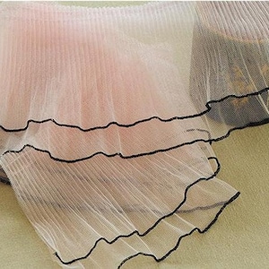 May include: Pink sheer fabric with black trim. The fabric is pleated and has a ruffled edge.