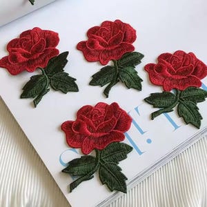 Op de afbeelding: Vier rode geborduurde rozenpatches met groene bladeren en stelen. De rozen zijn diep rood en de bladeren zijn donkergroen.