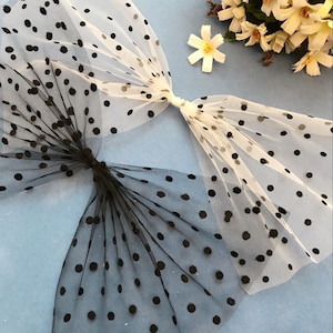 May include: Two pieces of black and white polka dot tulle fabric. The fabric is sheer and has a delicate texture. The fabric is arranged in a bow shape.